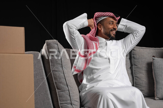 الانتقال إلى سكن جديد، صناديق وحاويات كرتونية، بورتريه لرجل عربي خليجي سعودي مبتسم يرتدي الزي التقليدي يجلس على كنبه رمادية بتعابير السرور، الترابط الأسري والحفاظ على الاستقرار العائلي، خلفيه سوداء