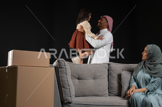 مفهوم الترابط الأسري، الانتقال الى منزل جديد, صناديق وحاويات كرتونية، بورتريه لسيده عربيه خليجيه سعوديه محجبة ترتدي العباءه الملونة تجلس على الاريكه الرماديه وتنظر الى زوجها وابنتها بحب، بناء علاقة أسرية ناجحة، مسؤولية المرأة تجاه أولادها، خلفيه سوداء