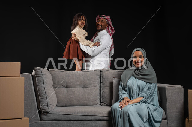 Building a successful and happy family relationship, moving to a new home, a portrait of a smiling Saudi Gulf Arab man wearing a traditional dress and shemagh carrying his daughter, cardboard boxes and containers, the concept of sitting and resting, black background