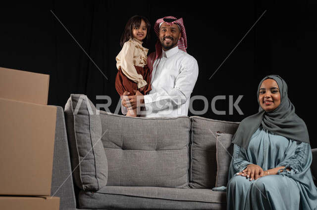 تدعيم أواصر المحبه والترابط الأسري، قضاء أوقات ممتعة مع الأبناء ,أجواء عائلية مليئة بالحب، صناديق وحاويات كرتونية، بورتريه لعائله عربيه سعوديه خليجيه تنظر الى الكاميرا بتعابير الفرح و السعادة، مفهوم الاستقرار العائلي وامتلاك سكن جديد, خلفيه سوداء