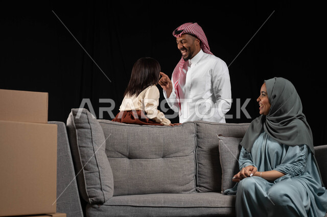 Cardboard boxes and containers, the concept of family stability and owning a new home, spending enjoyable times and creating strong bonds with children, portrait of a veiled Saudi Arabian Gulf woman wearing a colorful abaya, sitting on a gray sofa and looking at her husband and daughter with gestures of happiness and love, black background.
