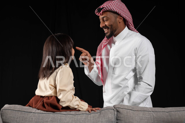 الاستمتاع والسرور بالجلسات العائليه الهادئه، بورتريه لرجل عربي خليجي سعودي يرتدي الزي التقليدي يقوم بمداعبه ابنته، قضاء أوقات ممتعه بايماءات السعاده مع الأبناء, الجلوس على الاريكه في غرفه المعيشة، الرعاية والاهتمام بالأطفال, خلفيه سوداء