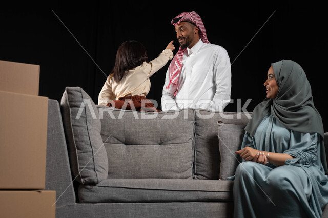 مفهوم الاستقرار العائلي وامتلاك سكن جديد، قضاء أوقات ممتعه وإنشاء روابط قويه مع الأبناء، صناديق وحاويات كرتونية، بورتريه لامراه عربيه خليجيه سعوديه محجبة ترتدي العباءة الملونة تجلس على كنبة رمادية وتنظر الى زوجها وابنتها بايماءات السعاده والمحبة، خلفيه سوداء