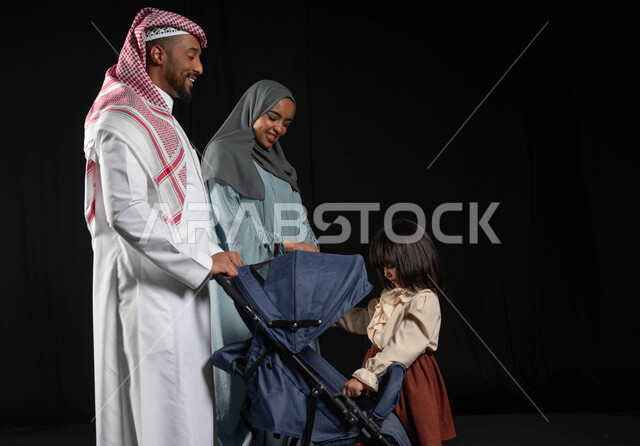 أجواء عائلية مليئة بالحب ,الاهتمام بالاطفال ورعايتهم، مفهوم الأسرة السعيدة المترابطة، بورتريه لفتاه عربيه خليجيه سعوديه ترتدي الزي الكاجوال تقف بجانب والديها وتشير باصبع السبابة إلى عربه الطفل، قضاء أوقات ممتعة مع الأبناء في الخارج ,خلفيه سوداء