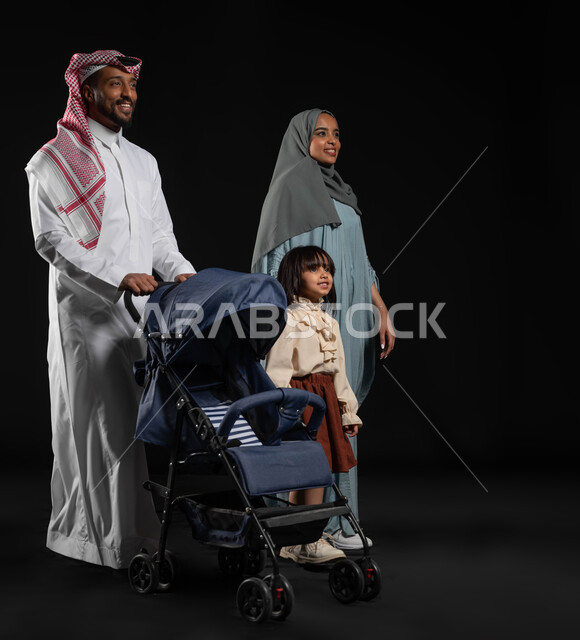 مفهوم الاسرة السعيدة المترابطة ,قضاء أوقات ممتعة مع الاطفال في الخارج ,المشي و التنزه مع العائلة، بورتريه لرجل عربي خليجي سعودي يرتدي الزي التقليدي يقوم بجر عربة الطفل برفقة زوجته وابنتهما، صوره الجسم بالطول الكامل، خلفيه سوداء