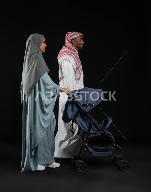 قضاء أوقات ممتعة مع الاطفال في الخارج ,المشي و التنزه مع العائلة، بورتريه من الجانب لامراه عربيه خليجيه سعوديه محجبة ترتدي العباءه الملونة  تقوم بجر عربة الطفل برفقة زوجها، الاهتمام بالابناء ورعايتهم، صوره الجسم بالطول الكامل، خلفيه سوداء