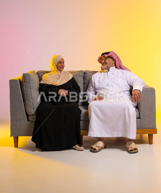 الألفة والمودة بين الأفراد في الأسر السعودية، بورتريه  لزوجين عربيين خليجيين سعوديين يرتديان الزي التقليدي يتبادلان أطراف الحديث بانسجام، قضاء أوقات ممتعة مع الزوجة، الجلوس على الأريكة بحب وحنان، صورة لكامل الجسم، خلفية ملونة