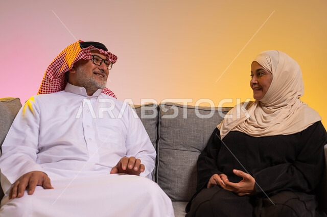 تبادل أطراف الحديث بين الزوجين بحب وانسجام، الألفة والمودة بين الأفراد في الأسر السعودية، بورتريه  لزوجين عربيين خليجيين سعوديين يرتديان الزي التقليدي يجلسان على الأريكة، قضاء أوقات ممتعة مع الزوجة، صورة لكامل الجسم، خلفية ملونة