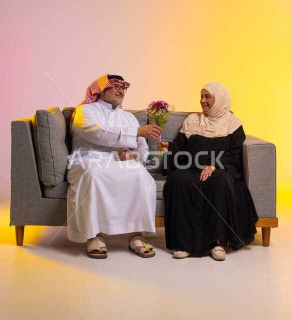 الأسرة السعودية السعيدة، تقدير واحترام الزوجة ومكافأتها، بورتريه لرجل كبير في السن عربي خليجي سعودي يرتدي الثوب التقليدي والشماغ يقوم بتقديم باقة ورد لزوجته، الجلوس على الأريكة بحب وحنان، تقديم الزهور في الاحتفالات والمناسبات، صورة لكامل الجسم، خلفية ملونة