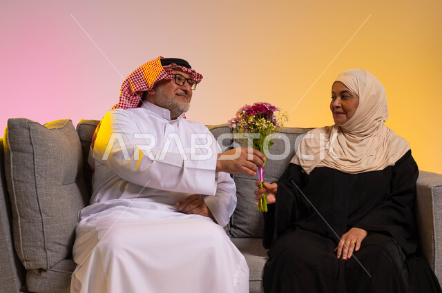 تقدير واحترام الزوجة ومكافأتها، بورتريه لرجل كبير في السن عربي خليجي سعودي يرتدي الثوب التقليدي والشماغ يقوم بتقديم باقة ورد لزوجته، الأسرة السعودية السعيدة، الجلوس على الأريكة بحب وحنان، تقديم الزهور في الاحتفالات والمناسبات، خلفية ملونة