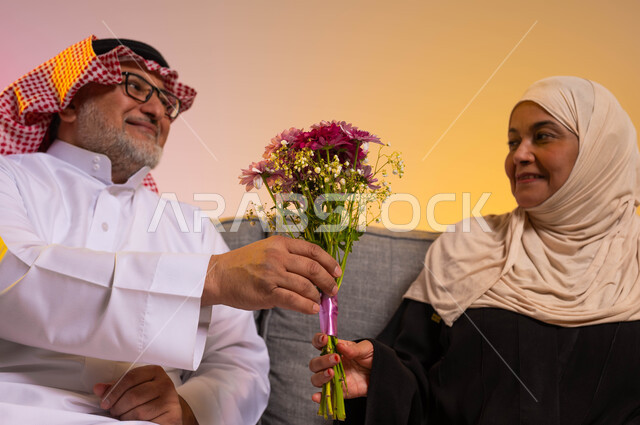 تقديم الزهور في الاحتفالات والمناسبات، الأسرة السعودية السعيدة، تقدير واحترام الزوجة ومكافأتها، بورتريه مقرب لرجل كبير في السن عربي خليجي سعودي يرتدي الثوب التقليدي والشماغ يقوم بتقديم باقة ورد لزوجته، الجلوس على الأريكة بحب وحنان، خلفية ملونة
