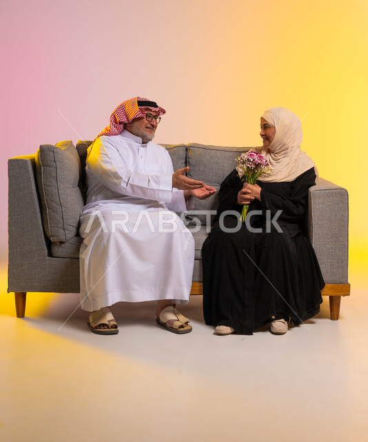 إيماءات الحنان والحب، مفهوم المودة والرحمة بين الزوجين، تبادل اطراف الحديث بين الزوجين، مفاجأة الزوج بالأزهار في الأعياد والمناسبات، بورتريه لرجل مسن عربي خليجي سعودي يمد يديه لزوجته بحب وتقدير، صورة لكامل الجسم، خلفية ملونة