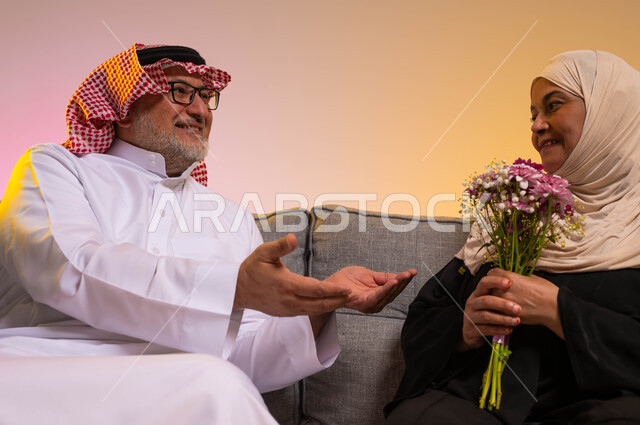 تبادل اطراف الحديث بين الزوجين، مفاجأة الزوج بالأزهار في الأعياد والمناسبات، إيماءات الحنان والحب، بورتريه لرجل مسن عربي خليجي سعودي يمد يديه لزوجته بحب وتقدير، مفهوم المودة والرحمة بين الزوجين، خلفية ملونة