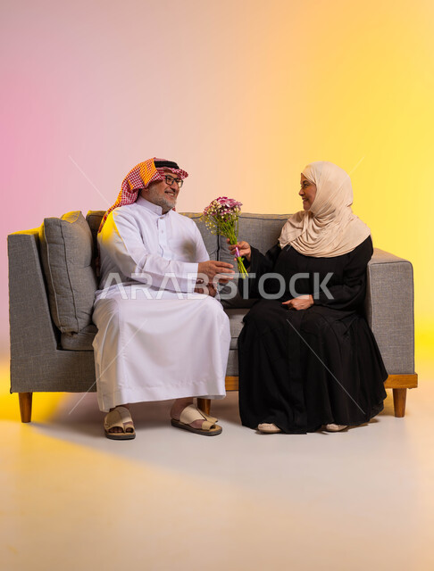 مفاجأة الزوج بالأزهار في الأعياد والمناسبات، إيماءات الحب والتقدير، الألفة والمودة بين أفراد الأسر السعودية، بورتريه لزوجين عربيين سعوديين يرتديان الزي التقليدي يجلسان على الأريكة يحتلان بعيد زواجهما، صورة لكامل الجسم، خلفية ملونة