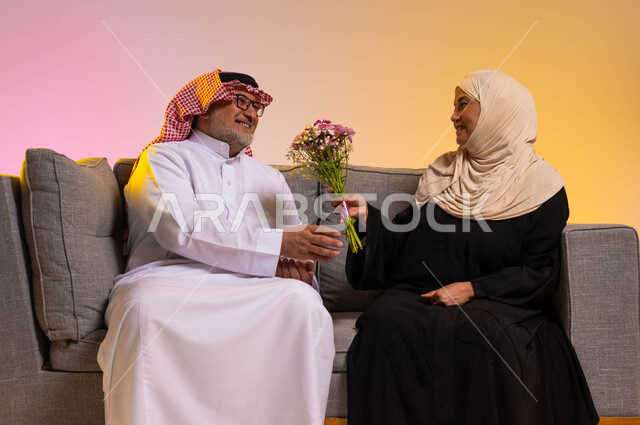 الاحتفال بعيد الزواج، الألفة والمودة بين أفراد الأسر السعودية، مفاجأة الزوج بالأزهار في الأعياد والمناسبات، إيماءات الحب والتقدير، بورتريه لزوجين عربيين سعوديين يرتديان الزي التقليدي يجلسان على الأريكة، خلفية ملونة