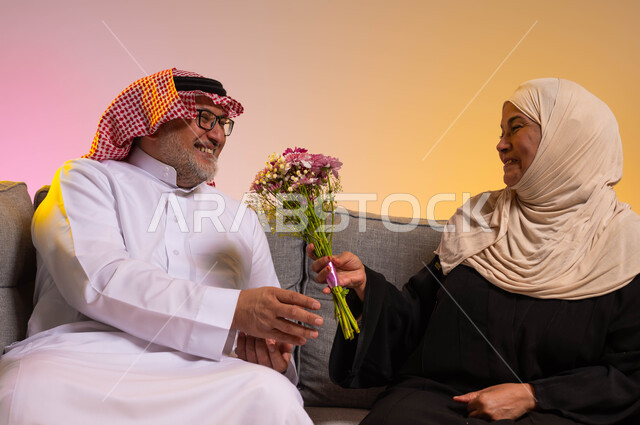 تعابير تدل على الحب والتقدير، الألفة والمودة بين الأفراد في الأسر السعودية، الاحتفال بعيد الزواج، مفاجأة الزوج بالأزهار في الأعياد والمناسبات، بورتريه لزوجين عربيين سعوديين يرتديان الزي التقليدي يجلسان على الأريكة، خلفية ملونة