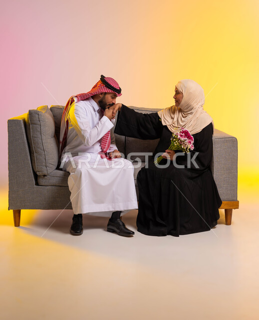 مفهوم بر الوالدين والإحسان إليهما، الترابط والتماسك في الأسر السعودية، التربية الصالحة للأبناء، تقديم الأزهار للأمهات في الأعياد والمناسبات، بورتريه لشاب عربي خليجي سعودي يجلس على الأريكة بجانب والدته يقوم بتقبيل يدها، صورة لكامل الجسم، خلفيه ملونة