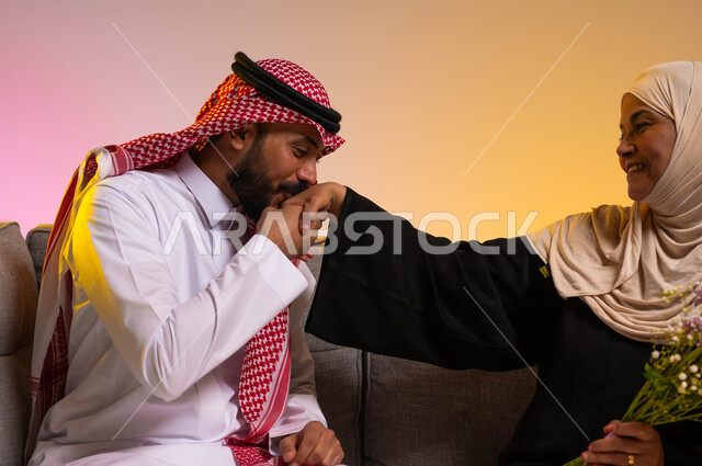 الاحترام والتقدير للأم ورد الجميل لها، الترابط والتماسك في الأسر السعودية، التربية الصالحة للأبناء، تقديم الأزهار للأمهات في الأعياد والمناسبات، بورتريه مقرب لشاب عربي خليجي سعودي يجلس على الأريكة بجانب والدته يقوم بتقبيل يدها، خلفيه ملونة