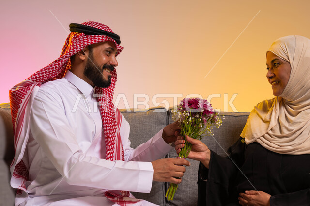 إهداء الأزهار في الأعياد والمناسبات، التربية الصالحة للأبناء، تماسك وترابط الأسرة السعودية، مفهوم بر الوالدين والإحسان إليهما، إظهار الحب والاحترام والتقدير للأم، بورتريه لشاب عربي خليجي سعودي يرتدي الزي التقليدي يجلس على الأريكة بجانب والدته، خلفية ملونة