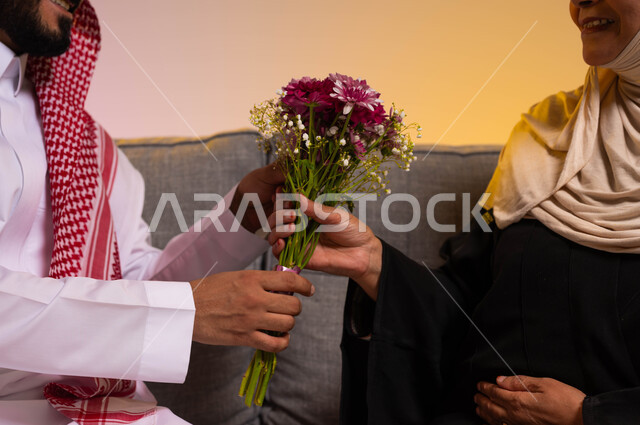 إدخال السرور إلى قلب الأم، التربية الصالحة للأبناء، تماسك وترابط الأسرة السعودية، مفهوم بر الوالدين والإحسان إليهما، إظهار الحب والاحترام والتقدير للأهل، بورتريه مقرب لأيدي شاب عربي خليجي سعودي يقوم بتقديم باقة من الأزهار لوالدته، خلفية ملونة