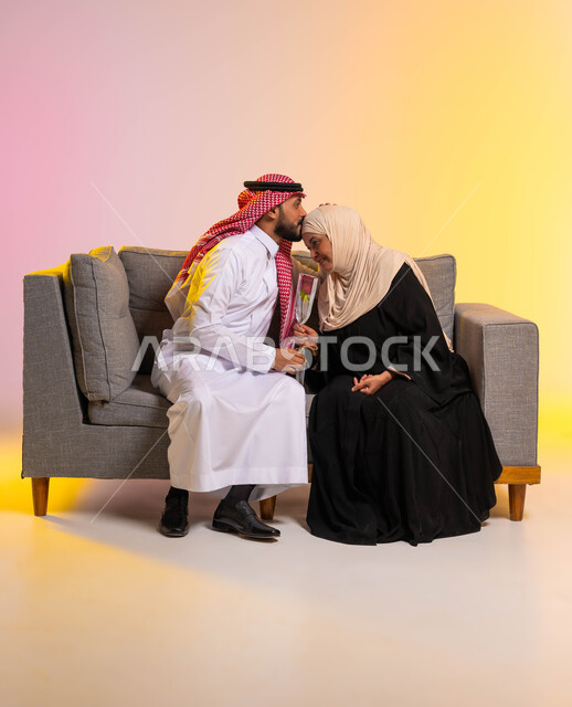 تماسك وترابط الأسرة السعودية، مفهوم بر الوالدين والإحسان إليهما، إظهار الحب والاحترام والتقدير للأم، بورتريه لشاب عربي خليجي سعودي يرتدي الزي التقليدي يجلس على الأريكة بجانب والدته يقوم بتقبيل رأسها، التربية الصالحة للأبناء، صورة لكامل الجسم، خلفية ملونة