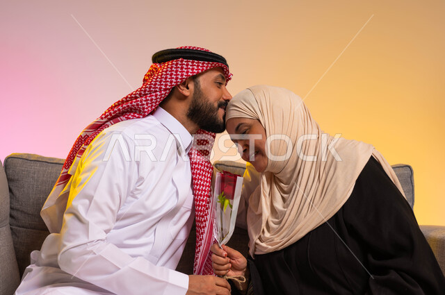 التربية الصالحة للأبناء، تماسك وترابط الأسرة السعودية، مفهوم بر الوالدين والإحسان إليهما، إظهار الحب والاحترام والتقدير للأم، بورتريه لشاب عربي خليجي سعودي يرتدي الزي التقليدي يجلس على الأريكة بجانب والدته يقوم بتقبيل رأسها، خلفية ملونة