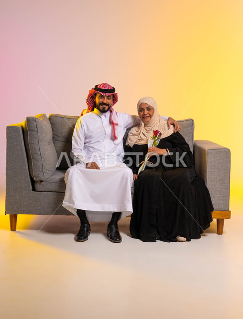 إظهار الحب والاحترام والتقدير للأم، النظر إلى الكاميرا بإيماءات الرضا والسرور، بورتريه لشاب عربي خليجي سعودي يرتدي الزي التقليدي يجلس على الأريكة بجانب والدته يقوم بمعانقتها، التربية الصالحة للأبناء، مفهوم بر الوالدين، صورة لكامل الجسم، خلفية ملونة
