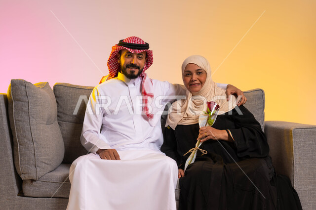 الاحتفال بعيد الأم، النظر إلى الكاميرا بإيماءات الرضا والسرور، بورتريه لشاب عربي خليجي سعودي يرتدي الزي التقليدي يجلس على الأريكة بجانب والدته يقوم بمعانقتها، إظهار الحب والاحترام والتقدير للأهل، التربية الصالحة للأبناء، مفهوم بر الوالدين، خلفية ملونة
