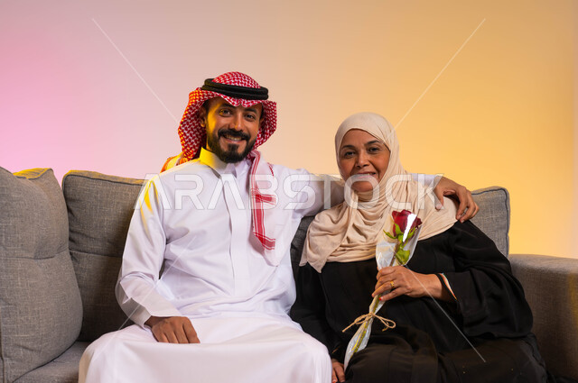 النظر إلى الكاميرا بإيماءات البهجة والسرور، بورتريه لشاب عربي خليجي سعودي يرتدي الزي التقليدي يجلس على الأريكة بجانب والدته يقوم بمعانقتها، إظهار الحب والاحترام للأم، التربية الصالحة للأبناء في الأسر السعودية، مفهوم بر الوالدين، خلفية ملونة