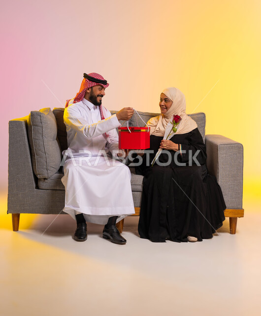 إحياء ذكرى عيد الأم، إيماءات الدهشة والسرور، مفاجأة الأم وإسعادها في الأعياد والمناسبات، بورتريه لشاب عربي خليجي سعودي يرتدي الزي التقليدي يجلس على الأريكة بجانب والدته يقوم بتقديم هدية لها، احترام الأهل وإظهار المحبة لهم، خلفية ملونة