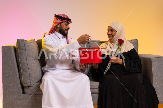 إيماءات الدهشة والسرور، مفاجأة الأم وإسعادها في الأعياد والمناسبات، إحياء ذكرى عيد الأم، بورتريه لشاب عربي خليجي سعودي يرتدي الزي التقليدي يجلس على الأريكة بجانب والدته يقوم بتقديم هدية لها، احترام الأهل وإظهار المحبة لهم، خلفية ملونة