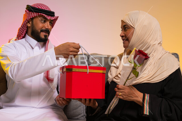 إحياء ذكرى عيد الأم، احترام الأهل وإظهار المحبة لهم، إيماءات الدهشة والسرور، مفاجأة الوالدين في الأعياد والمناسبات، بورتريه لشاب عربي خليجي سعودي يرتدي الزي التقليدي يجلس على الأريكة بجانب والدته يقوم بتقديم هدية لها، خلفية ملونة