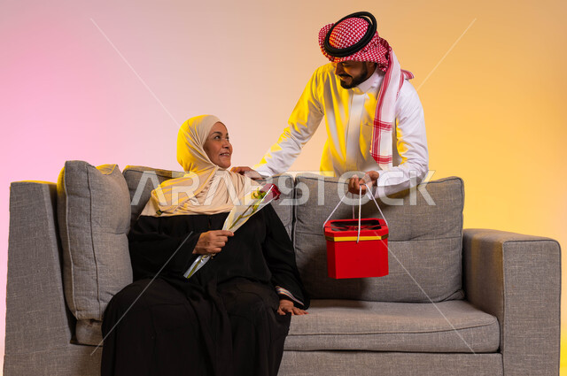 تبادل نظرات المودة بين الأهل، تقدير جهود الأمهات والإحسان إليهم، بورتريه مقرب لشاب عربي خليجي سعودي يرتدي الزي التقليدي يقف خلف والدته يقوم بتقديم صندوق هدايا لها، سيدة عربية تجلس على الأريكة تحمل بيدها وردة حمراء، الاحتفال بعيد الأم، خلفية ملونة