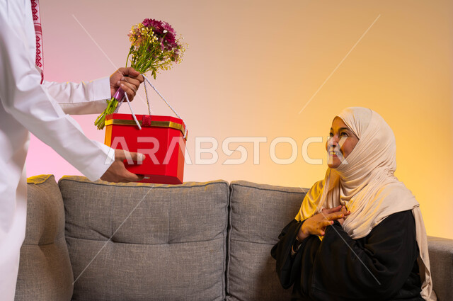 إعداد المفاجآت السعيدة في الأعياد، الاحتفال بعيد الأم، بورتريه لأيدي شاب عربي خليجي سعودي يقوم بتقديم صندوق أحمر مع باقة من الأزهار لوالدته، تعابير الدهشة والسرور، مفهوم بر الوالدين والإحسان إليهما، التربية الصالحة للأبناء في الأسر السعودية، خلفية ملونة