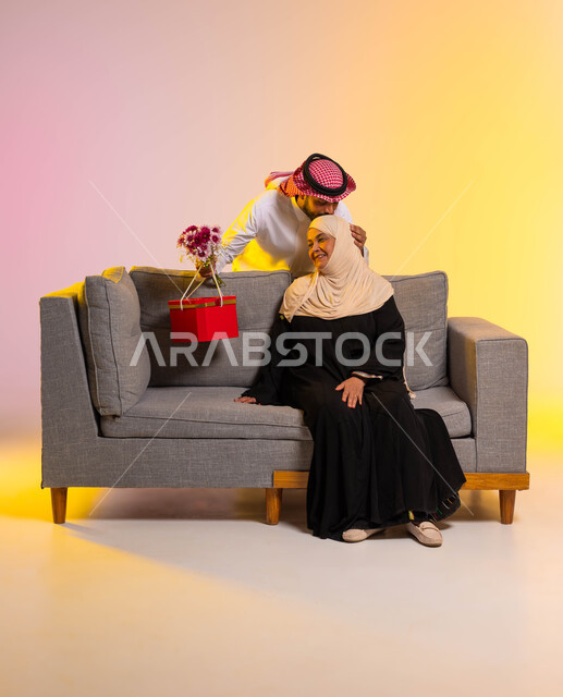 تقديم الهدايا في الأعياد والمناسبات، إيماءات الرضا والسعادة، إحياء ذكرى عيد الأم، مفهوم احترام وتقدير الوالدين ورد الجميل لهما، بوتريه لشاب عربي خليجي سعودي يقوم بتقبيل رأس والدته، إدخال السرور إلى قلب الأهل، تماسك الأسرة السعودية، صورة لكامل الجسم، خلفية ملونة