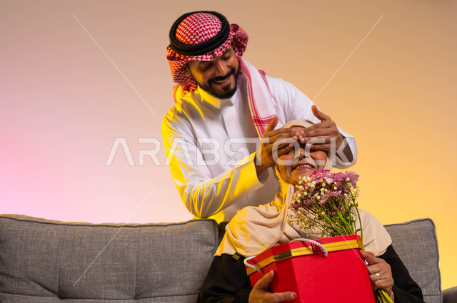 التربية الصالحة للأبناء، إدخال السرور إلى قلب الأهل، إسعاد الأم بالهدايا والأزهار في الأعياد والمناسبات، بورتريه مقرب لشاب عربي خليجي سعودي يرتدي الزي التقليدي يقوم بإغماض عيني والدته، المحبة والألفة بين أفراد الأسرة السعودية، خلفية ملونة