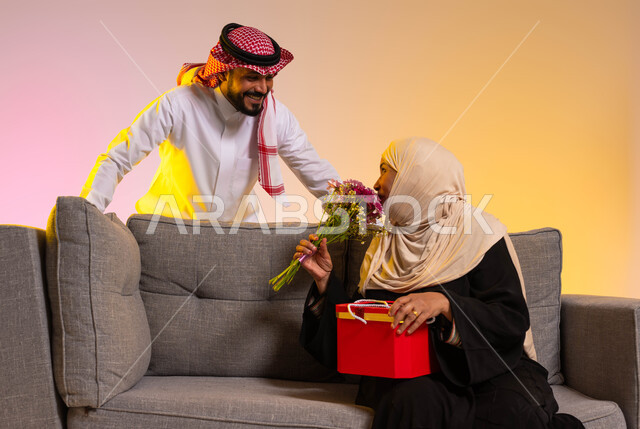 احترام وتقدير وبر الوالدين، تبادل نظرات المودة بين الأم والابن، بورتريه مقرب لشاب عربي خليجي سعودي يرتدي الثوب التقليدي والشماغ يقف خلف والدته، سيدة عربية تجلس على الأريكة تحمل باقة من الأزهار، الاحتفال بالأعياد والمناسبات العائلية، خلفية ملونة