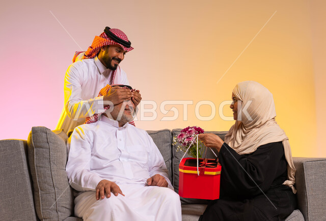 مفاجأة الزوج بالهدايا والأزهار في الأعياد، إدخال الفرح إلى قلب الأب، مفهوم بر الوالدين، بورتريه لزوجان عربييان خليجييان سعوديان يجلسان على الأريكة، شاب يقوم بإغماض عيني والده، الألفة والمحبة في الأسرة السعودية، إيماءات البهجة والسرور، خلفية ملونة