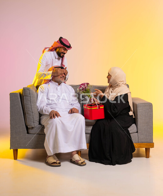 تبادل المودة والمحبة بين أفراد الأسرة، مفاجأة الزوج بالهدايا والأزهار في الأعياد، إدخال الفرح إلى قلب الأب، بورتريه لزوجان عربييان خليجييان سعوديان يجلسان على الأريكة، شاب يقوم بإغماض عيني والده، إيماءات البهجة والسرور، خلفية ملونة