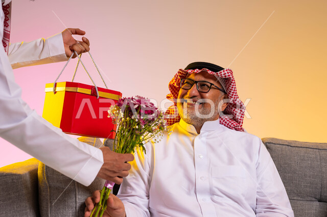 مفهوم بر الوالدين، الاحتفال بأعياد الميلاد، بورتريه مقرب لسيد عربي خليجي سعودي يرتدي الزي التقليدي يجلس على الأريكة، شاب يقوم بتقديم هدية مع باقة من الأزهار لوالده، الألفة والمحبة في الأسرة السعودية، إدخال الفرح إلى قلب الأب،إيماءات البهجة والسرور، خلفية ملونة