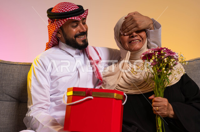 مفهوم المحبة والألفة بين أفراد العائلة، أهمية العائلة وإحسان للأمهات، إهداء الهدايا والأزهار في الأعياد والمناسبات السعيدة، بورتريه مقرب لشاب عربي خليجي سعودي يرتدي الثوب التقليدي يغمض عيني والدته لمفاجأتها بهدية عيد الأم، خلفية ملونة