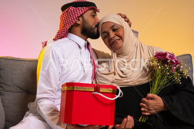 مفهوم الإحسان وتقدير الوالدة، بورتريه مقرب لشاب عربي خليجي سعودي يرتدي الثوب التقليدي والشماغ يقوم بتقبيل رأس والدته، إهداء الهدايا والزهور في الأعياد والمناسبات، التربية الصالحة للأبناء، إدخال الفرحة والسرور الى قلب الأم، خلفية ملونة