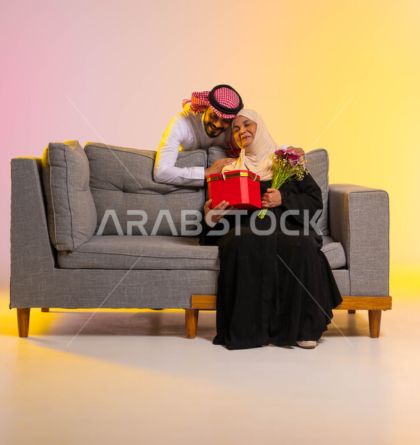 الأعياد والمناسبات السعيدة، مفاجأة الوالدة بهدية عيد الأم، أجواء عائلية مليئة بالطمأنينة والحب، بورتريه لامرأة عربية خليجية سعودية محجبة ترتدي عباءة تقليدية تجلس على أريكة رمادية وتحمل هدية وباقة ورود، محبة الأمهات والإحسان لهم، الطول الكامل للجسم، خلفية ملونة