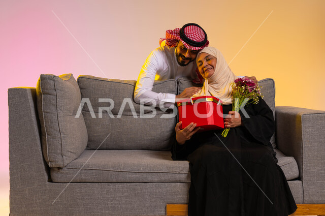 المناسبات والأعياد السعيدة، مفاجأة الوالدة بهدية عيد الأم، أجواء عائلية مليئة بالطمأنينة والحب، بورتريه لامرأة عربية خليجية سعودية محجبة ترتدي عباءة تقليدية تجلس على أريكة رمادية وتحمل هدية وباقة ورود، محبة الأمهات والإحسان لهم، خلفية ملونة