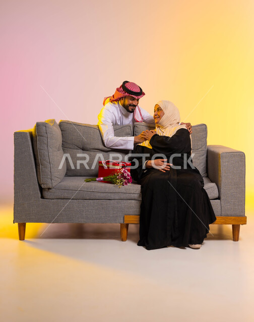 مفهوم رضى الأمهات وبر الوالدين، بورتريه لامرأة عربية خليجية سعودية محجبة ترتدي عباءة تقليدية تجلس على أريكة رمادية تقوم بالنظر الى ابنها بحب، شاب يفاجئ والدته بهدية وباقة ورد، إحياء ذكرى عيد الأم بالهدايا والمفاجآت، الطول الكامل للجسم، خلفية ملونة