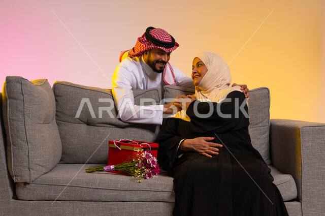 مفهوم بر الوالدين ورضى الأمهات، بورتريه لامرأة عربية خليجية سعودية محجبة ترتدي عباءة تقليدية تجلس على أريكة رمادية تقوم بالنظر الى ابنها بحب، شاب يفاجئ والدته بهدية وباقة ورد، إحياء ذكرى عيد الأم بالهدايا والمفاجآت، خلفية ملونة