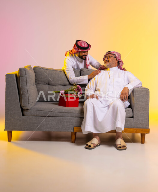 تقدير واحترام وبر الوالدين، تبادل نظرات المحبة والألفة بين الأب والابن، بورتريه لرجل عربي خليجي سعودي يجلس على أريكة رمادية، شاب يفاجئ والده بهدية وباقة ورد، ارتداء الأزياء التقليدية في المناسبات والأعياد السعيدة، الطول الكامل للجسم، خلفية ملونة