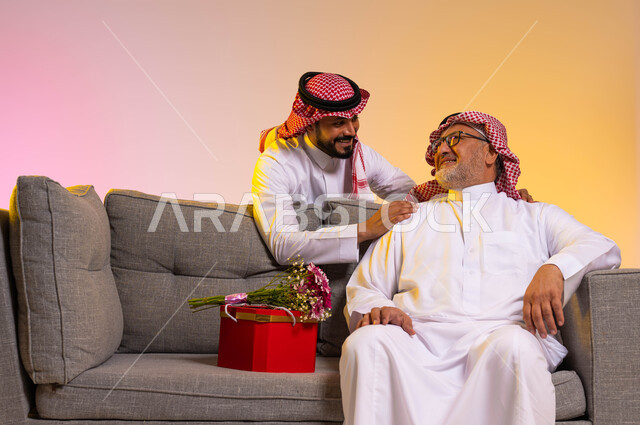 Wearing traditional costumes on happy occasions and holidays, exchanging looks of love and affection between father and son, a portrait of a Saudi Gulf Arab man sitting on a gray sofa, a young man surprising his father with a gift and a bouquet of flowers, appreciation and respect for filial piety, a colorful background.