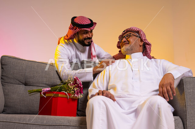 تبادل نظرات المحبة والألفة بين الأب والابن، بورتريه لرجل عربي خليجي سعودي يجلس على أريكة رمادية، شاب يفاجئ والده بهدية وباقة ورد، ارتداء الأزياء التقليدية في المناسبات والأعياد السعيدة، تقدير واحترام وبر الوالدين، خلفية ملونة