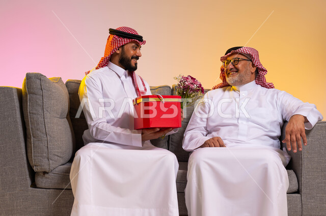 تعابير البهجة والسرور، مفهوم المحبة والألفة بين أفراد العائلة، بورتريه لرجل عربي خليجي سعودي يرتدي الزي التقليدي والشماغ يقوم بالنظر الى ابنه بحب، شاب يقدم هدية وباقة ورد لوالده بمناسبة عيد ميلاده، الاحتفال بالأعياد والمناسبات، خلفية ملونة